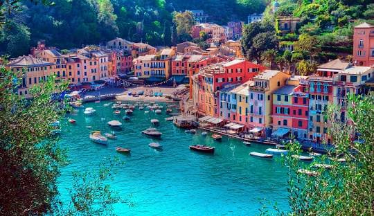 Colorful waterfront village with pastel buildings and turquoise water dotted with boats, surrounded by green hillside vegetat